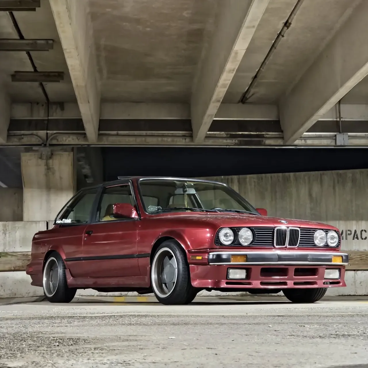 A burgundy BMW E30 under a concrete overpass, seen from the front three-quarter